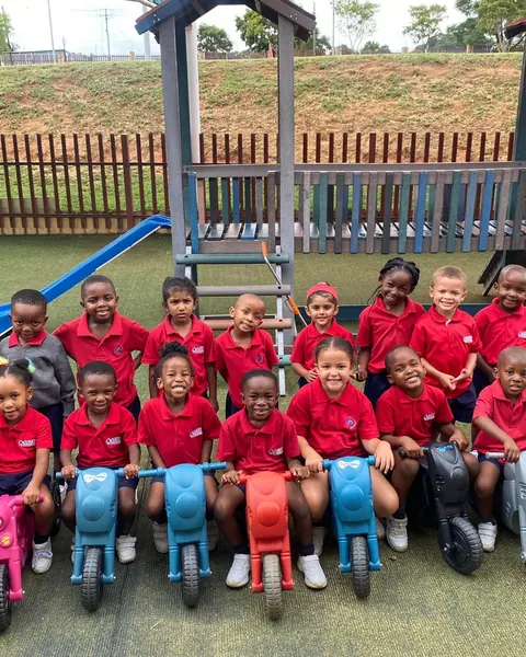 A Grade R class in red shirts sitting on colourful balance bikes by the jungle gym